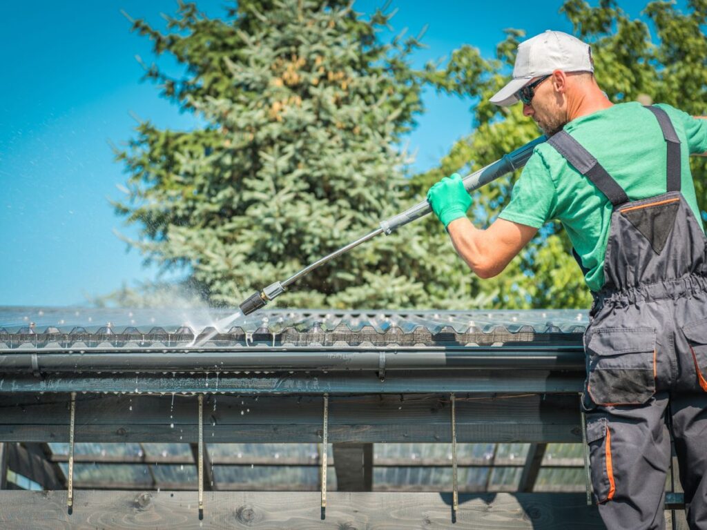 Worker pressure washing a metal gutter under a clear sky.