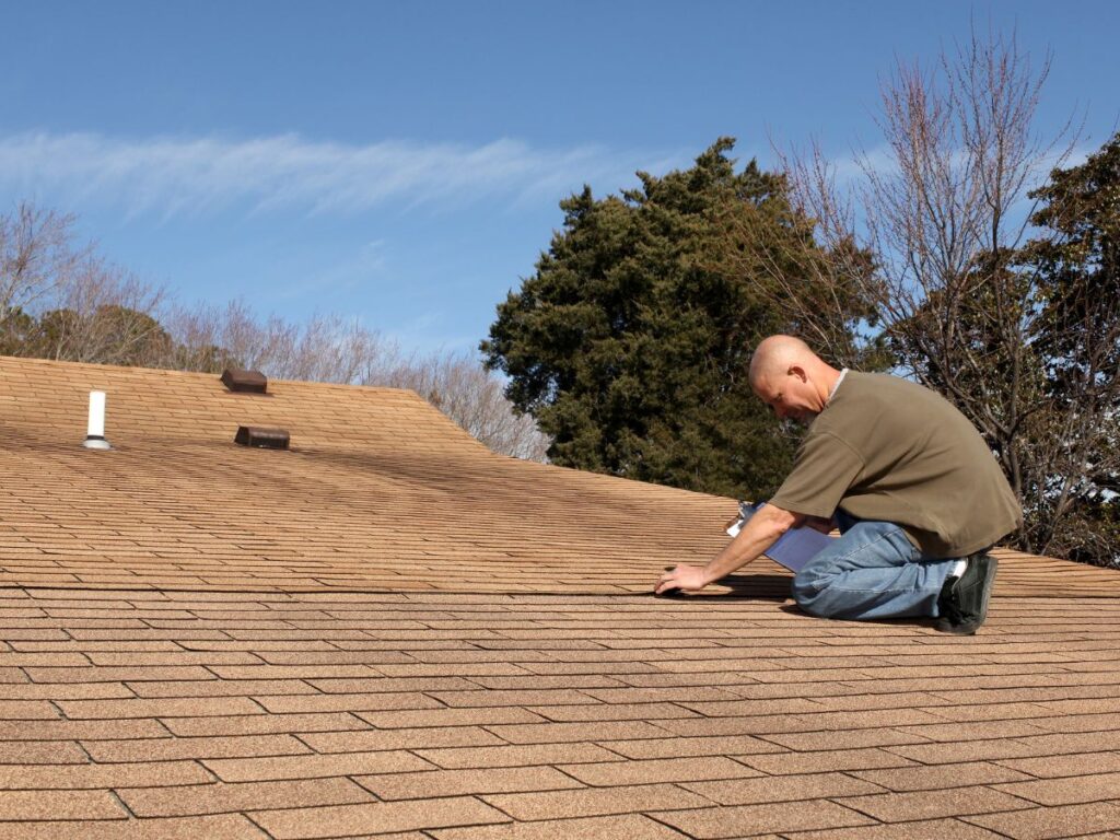 Man inspecting shingles on a roof under a clear blue sky.