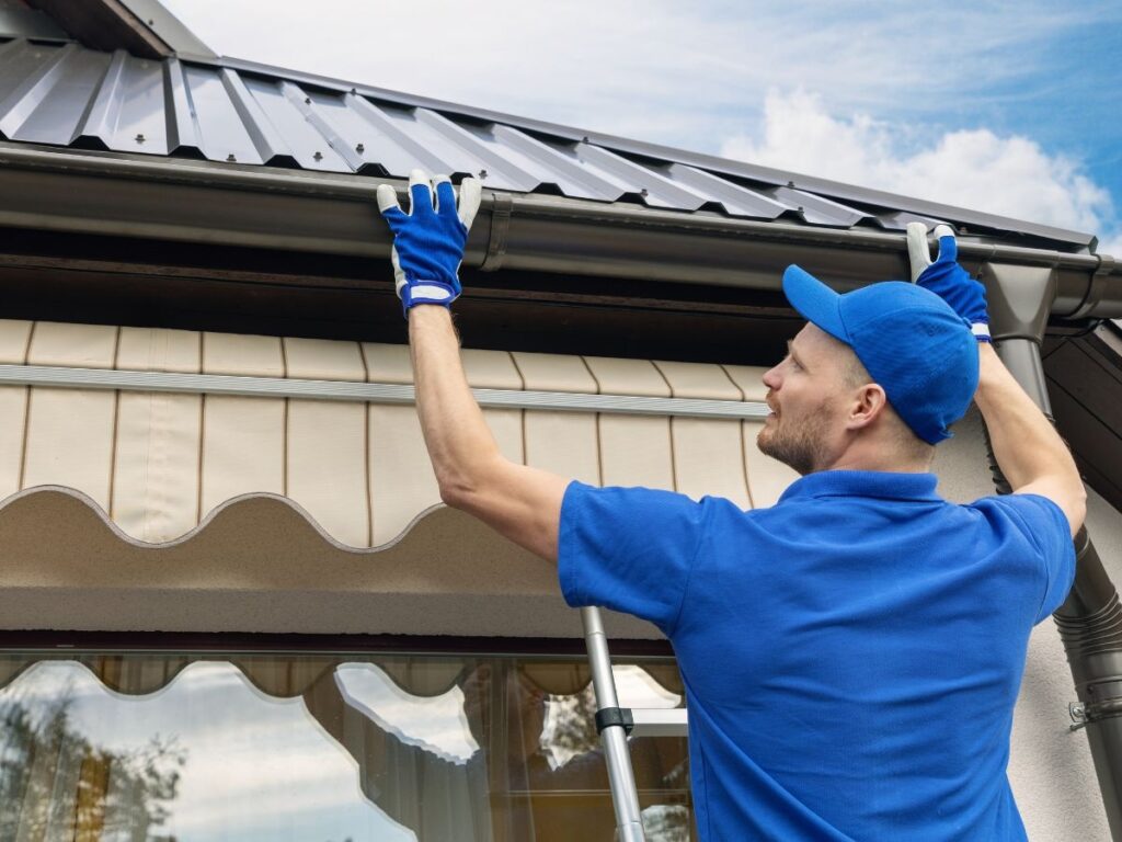 A man wearing a blue hat and glove trying to repair the roof gutter.